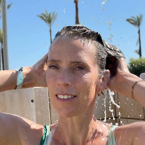 A woman outdoors under running water, washing her hair. She is wearing a light blue wristwatch and gold hoop earrings, with palm trees in the background.