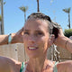 A woman outdoors under running water, washing her hair. She is wearing a light blue wristwatch and gold hoop earrings, with palm trees in the background.