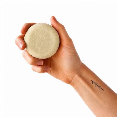 A hand holding a round, pale beige soap bar against a solid white background.
