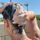 A close-up lifestyle shot of a person washing their hair outdoors, with soapy lather covering the dark hair and hands, set against a background of blue sky and palm trees.