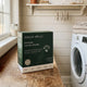 A box of Wash Wild Everyday Laundry Powder sits on a wooden counter in a bright laundry room next to a white washing machine.