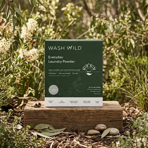 Wash Wild Everyday Laundry Powder box standing on a wooden log outdoors among green plants and small flowers.