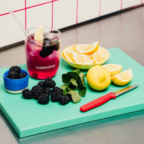 Glass of waterdrop microdrink blackberry with lemon slices, blackberries, and mint leaves on a green cutting board.