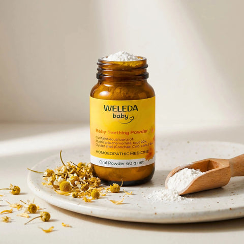 Weleda Baby Teething Powder on a plate sprinkled with chamomile flowers and a mini wooden scoop of powder.