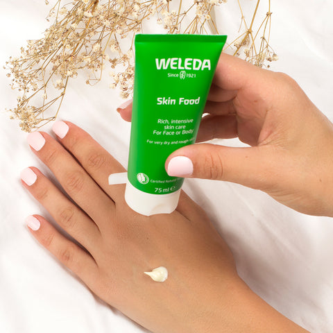 Hand holding a Weleda Skin Food tube with a small amount of cream on the palm against a white background.