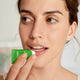 A close-up of a woman applying a green tube of skin food nourishing lip balm to her lips, with a glossy finish.