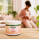 Weleda Stretch Mark Body Butter 150ml jar on a table with a blurred background of a woman sitting on a couch.