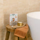 A box of zazen Infinity Bath Tablets sits on a rustic wooden stool next to a white freestanding bathtub, with a terracotta bath towel and a gold tin candle resting on it.